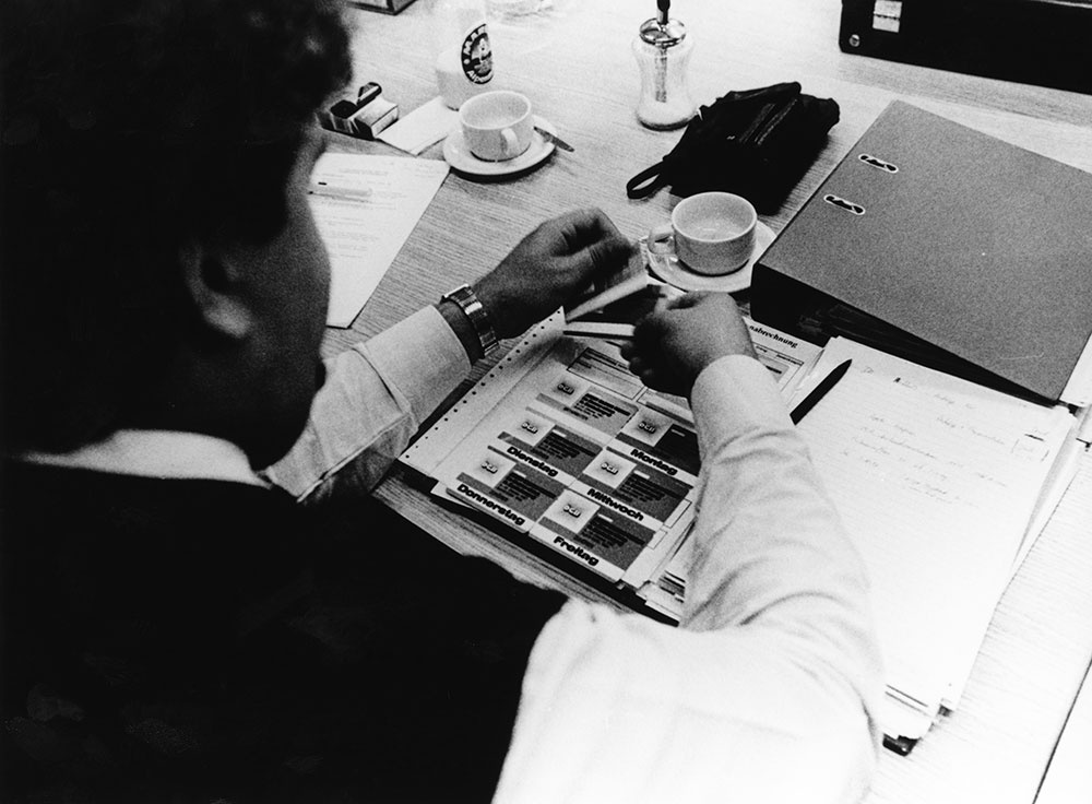 Man sieht über die Schulter einer sitzenden Person auf einen Tisch, darauf stehen Kaffeetassen, ein Ordner und eine geöffnete Mappe mit Marken, auf denen ÖGB und die Wochentage steht. © IHSF-Archiv