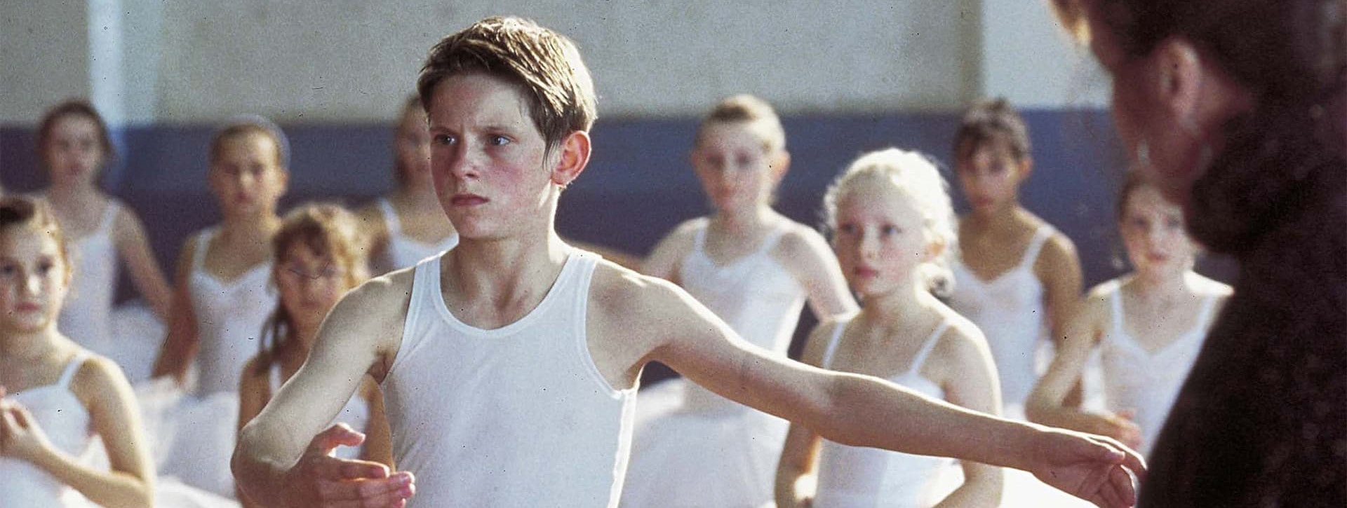 Im Fokus des Bildes steht ein angestrengt wirkender Junge in Sportkleidung, um ihn herum Mädchen in Ballettkleidung, am rechten Bildrand sieht man eine erwachsene Frau. © https://www.salamontjuic.org/en/movie/billy-elliot/