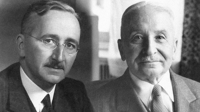 Beide tragen Anzüge, weiße Hemden und Krawatten. Sie haben Schnurbärte und ihr Haar nach hinten gekämmt.  Hayek trägt eine randlose Brille. © wikipedia