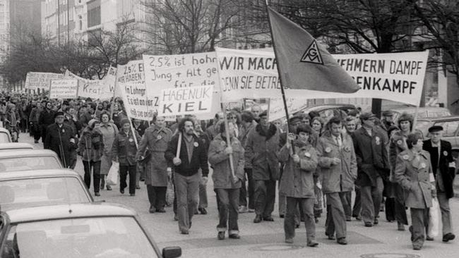  Protestzug der Hagenuk-Mitarbeiter in der Gneisenaustraße. Links neben der IG Metall-Fahne der Erste Bevollmächtigte der IG Metall Kiel Alfred Prezewowsky, dann Harald Carlsen, Betriebsrat bei Hagenuk. Ganz vorn links Betriebsrat Karlheinz Göttsche (links) und rechts der Vorsitzende des Hagenuk-Betriebsrats Günter Schmollmann. © Wikimedia / Stadtarchiv Kiel