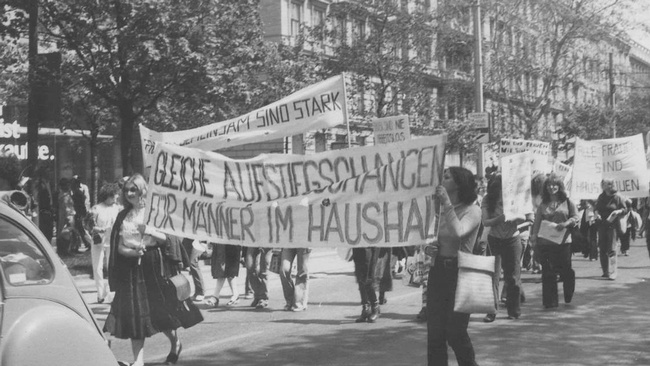 Ein Demonstrationszug, Frauen tragen Banner, auf dem vorne steht "Gleiche Aufstiegschancen f&uuml;r M&auml;nner im Haushalt", weiter hinten "Alle Frauen sind Hausfrauen". &copy; Burgi Hirsch, Kreisky-Archiv