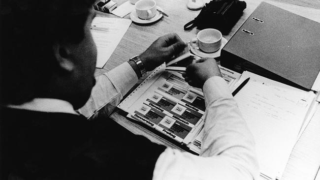 Man sieht über die Schulter einer sitzenden Person auf einen Tisch, darauf stehen Kaffeetassen, ein Ordner und eine geöffnete Mappe mit Marken, auf denen ÖGB und die Wochentage steht. © IHSF-Archiv