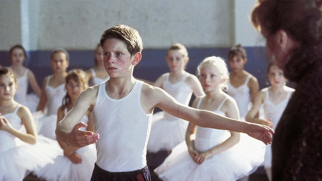 Im Fokus des Bildes steht ein angestrengt wirkender Junge in Sportkleidung, um ihn herum Mädchen in Ballettkleidung, am rechten Bildrand sieht man eine erwachsene Frau. © https://www.salamontjuic.org/en/movie/billy-elliot/