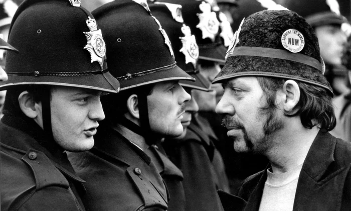 Auf der linken Seite stehen eine Reihe Polizisten. Rechts, nahe vor ihnen, steht ein einzelner Mann, der dem Polizisten vor ihm mutig in die Augen schaut. Auf seiner Mütze ist ein Button, auf dem steht Support the Miners. NUM. Stop Pit Closures © The Guardian / Eyevine / picturedesk.com